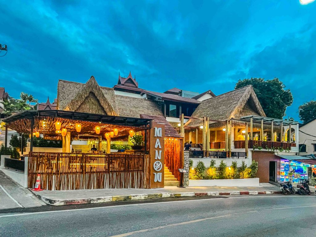 Evening exterior of Manow Brunch & Bistro in Kata Karon with thatched roof, glowing lanterns, and rustic design.