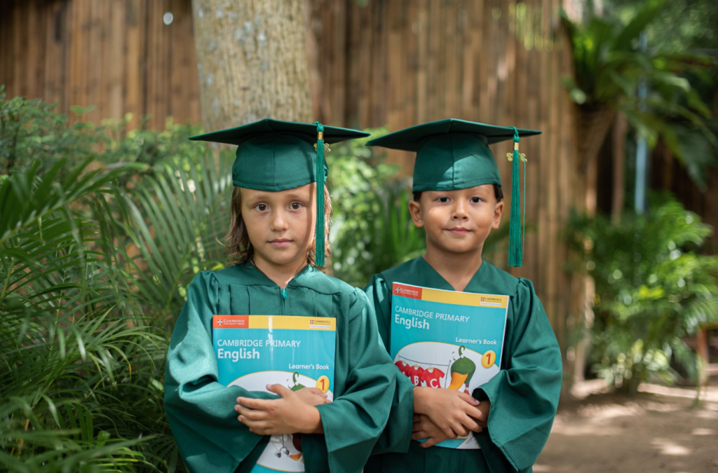 Two young students in green graduation gowns holding Cambridge Primary English books at an international school in Phuket